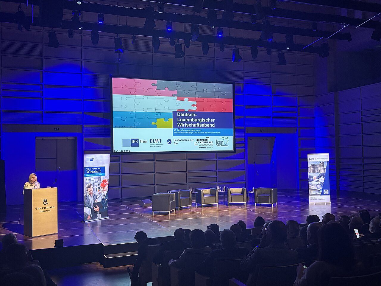 Eine Frau steht hinter einem Podium neben einem Screen, auf dem der Titel ‚Deutsch-Luxemburgischer Wirtschaftsabend‘ angezeigt wird. Davor sind Ledersessel für die Gastsprecher des Abends aufgestellt.