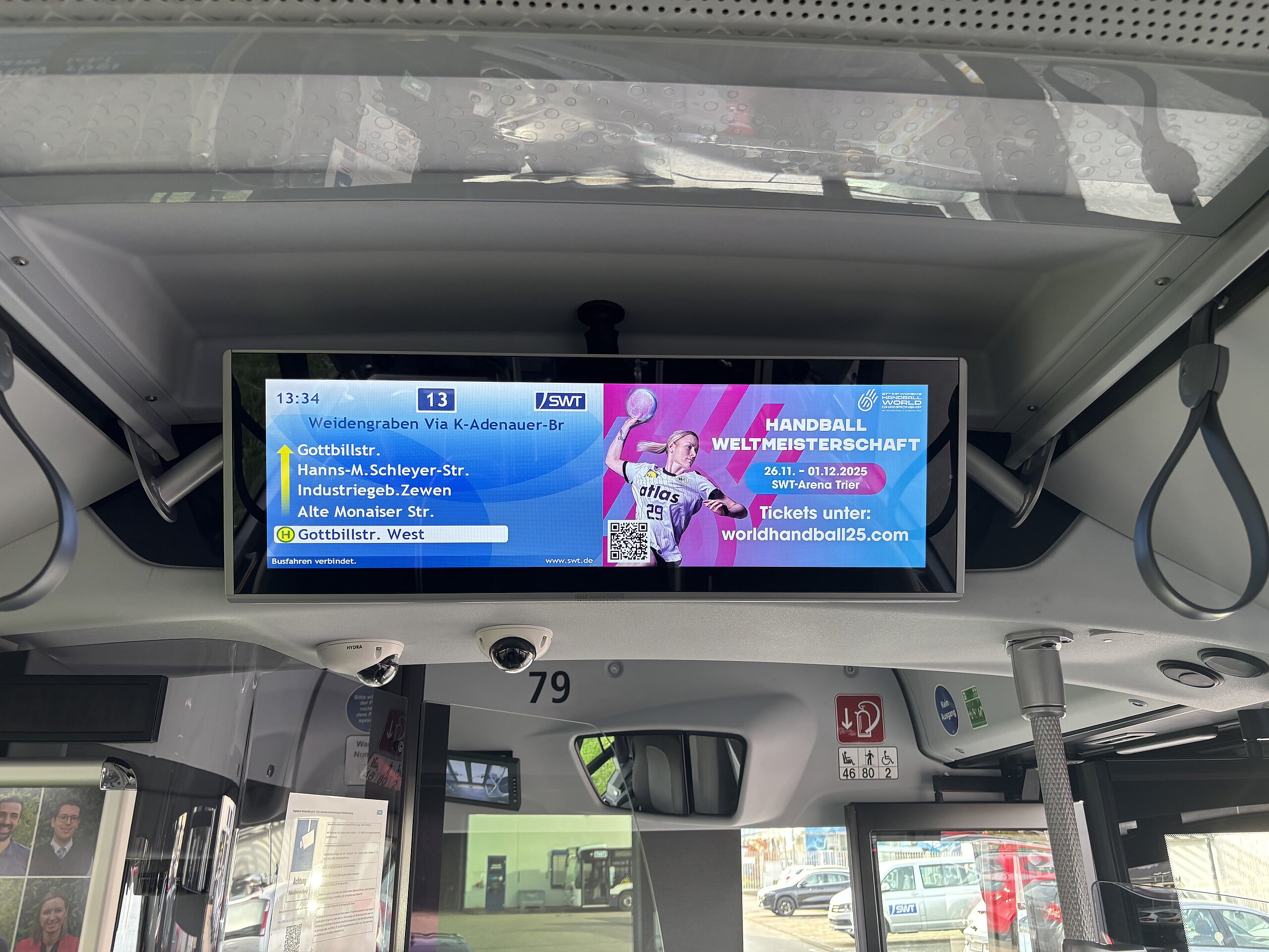 Das Innere eines Stadtbusses mit digitalem Werbescreen im Fahrgastbereich, der eine Anzeige für die Handball-Weltmeisterschaft der Damen zeigt.