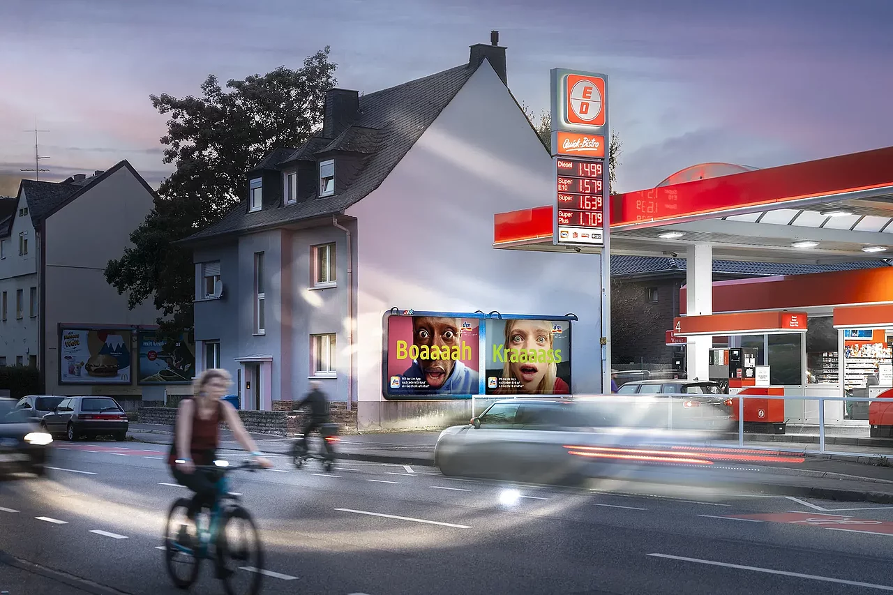 Großflächenplakate an einer Hauswand neben einer Tankstelle bei Abenddämmerung, Autos und Radfahrer auf der Straße.
