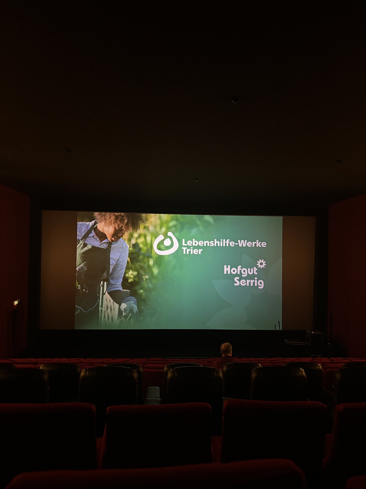 Leerer Kinosaal mit beleuchteter Leinwand, auf der ein Motiv des Hofgut Serrig zu sehen ist. Das Motiv zeigt eine Frau mit grüner Schürze im Garten sowie die Aufschrift „Lebenshilfe-Werke Trier“.