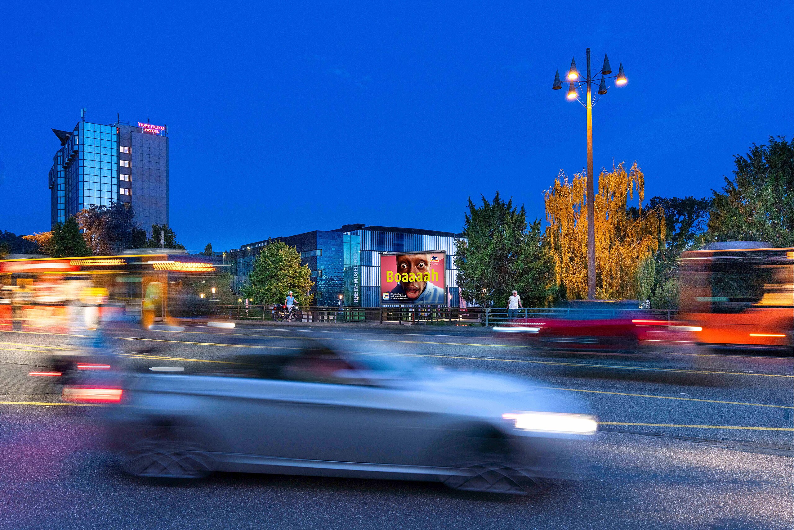 Großplakat mit farbenfrohem „Boaaah“-Motiv an einer vielbefahrenen Straße bei Nacht, vorbeifahrende Autos und Busse – buchbar über Plakat-verkauft.de