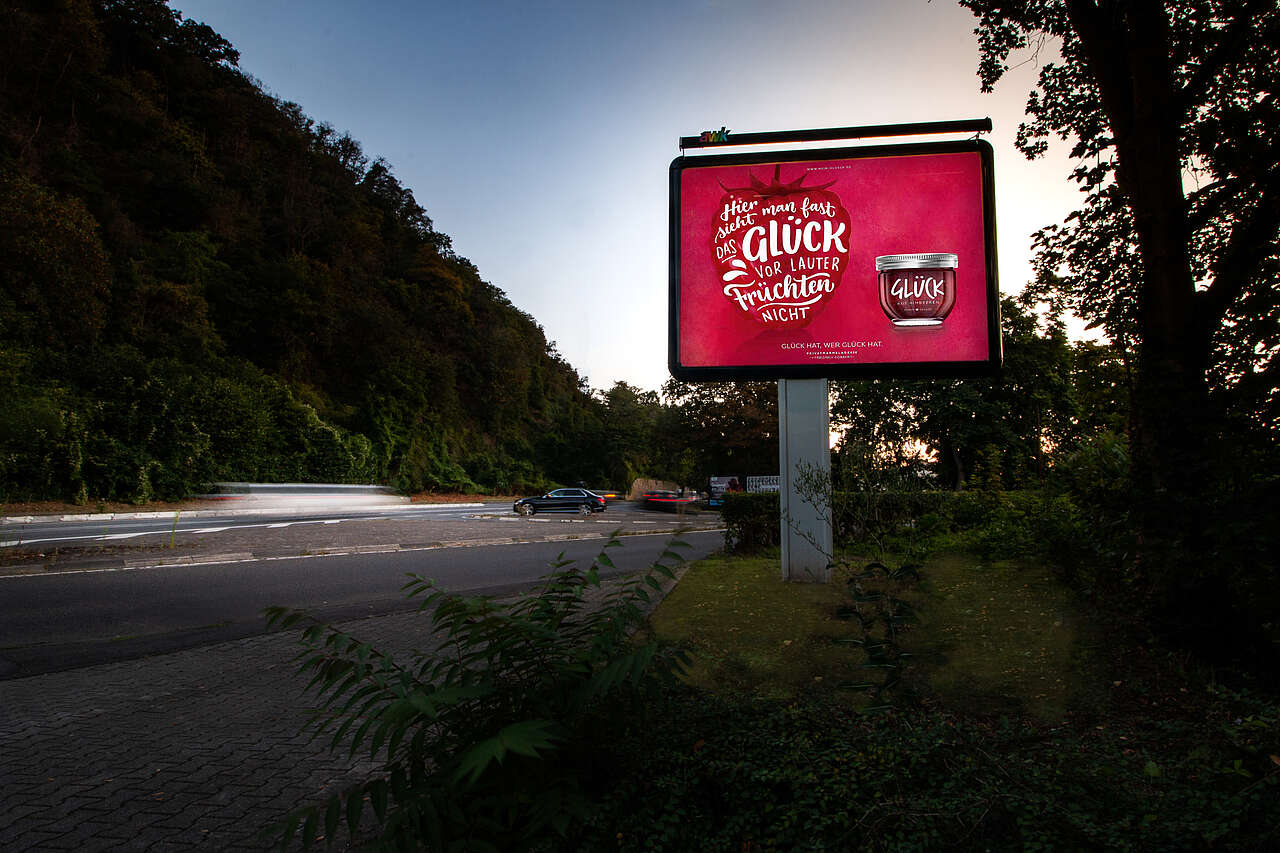 plakat-verkauft.de_megalight_koblenz-glueck Citystar an befahrener Straße am Waldrand mit Motiv von Glück