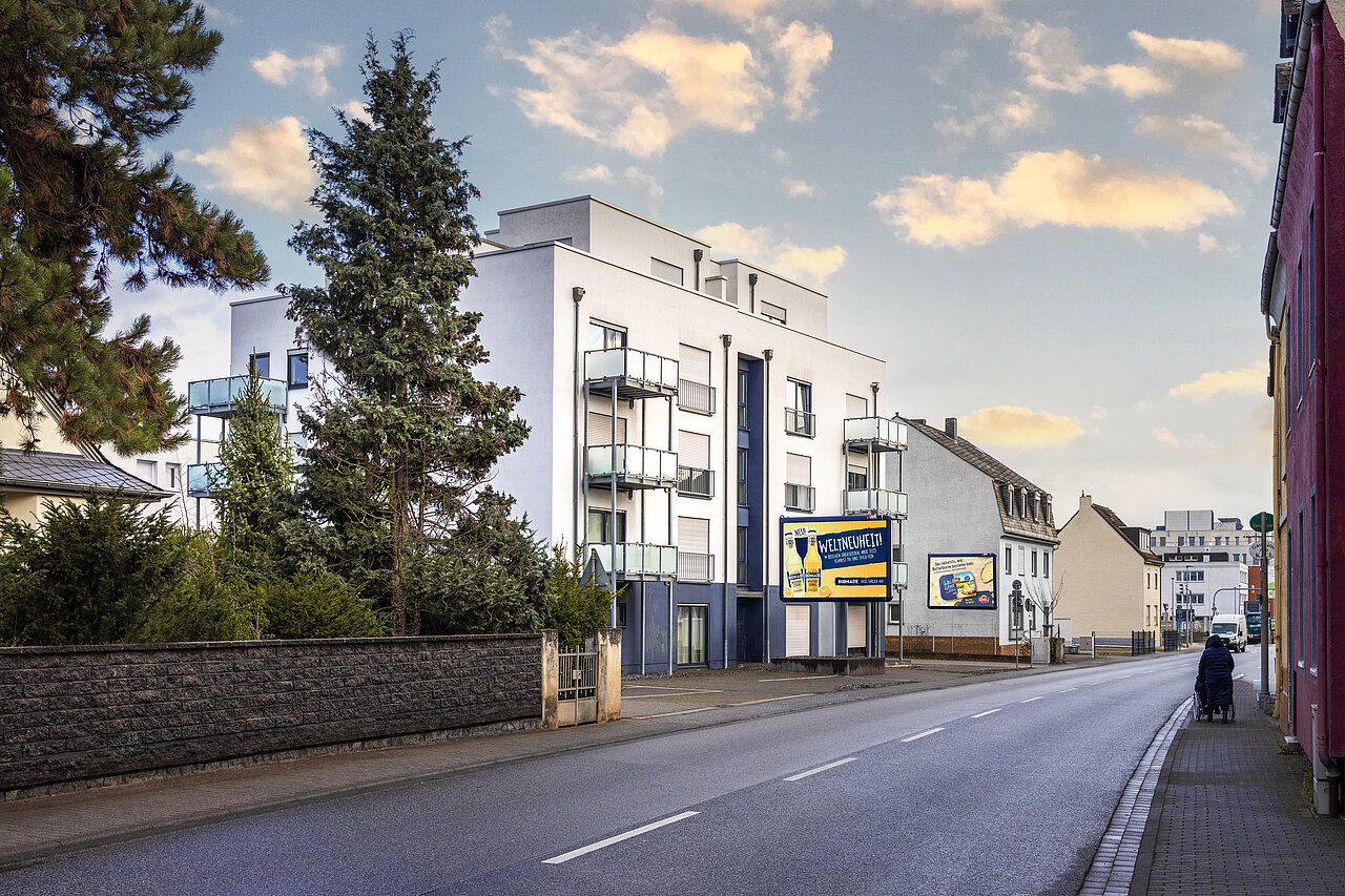 Eine Großfläche, montiert auf dem Vorgarten eines modernen Mehrfamilienhauses, welches sich an einer gut befahrenen Straße befindet.