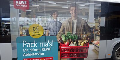 Traffic Board auf einem Bus für REWE mit dem Abholservice, zeigt einen Kunden mit einer Einkaufskiste und dem Slogan „Pack ma’s!“.