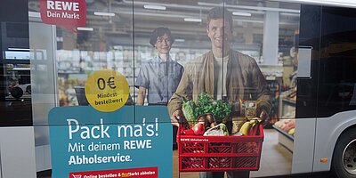 Traffic Board auf einem Bus für REWE mit dem Abholservice, zeigt einen Kunden mit einer Einkaufskiste und dem Slogan „Pack ma’s!“.