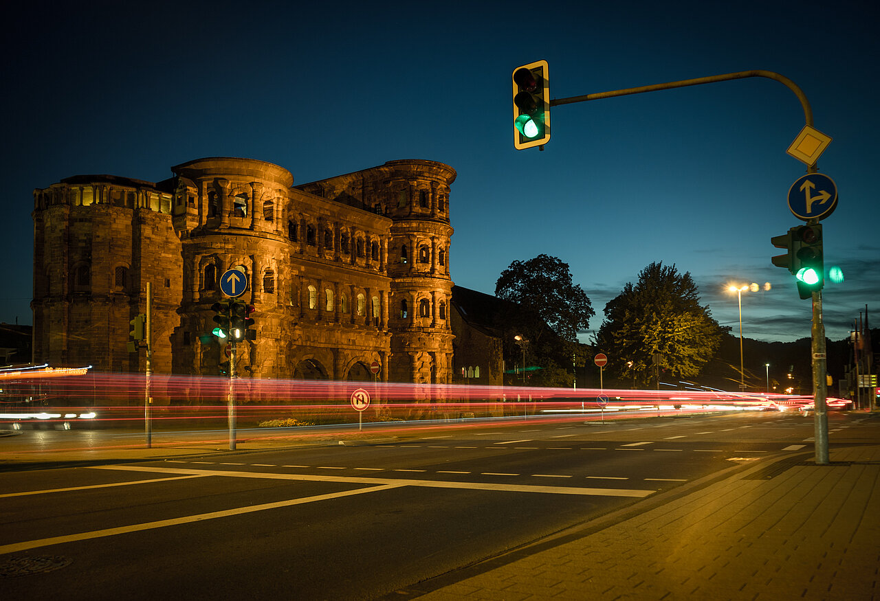 Blick auf eine Straßen-Kreuzung neben der Porta Nigra bei Nacht in der deutschen Stadt Trier in Rheinland-Pfalz. 