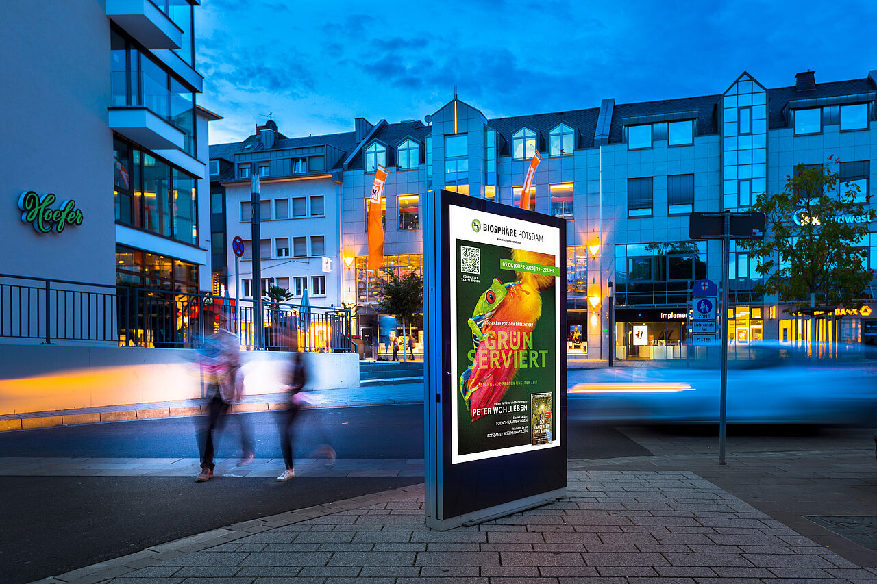 Beleuchtetes Citylight-Poster in der Potsdamer Innenstadt mit Werbemotiv der Biosphäre Potsdam, Passanten im Hintergrund.
