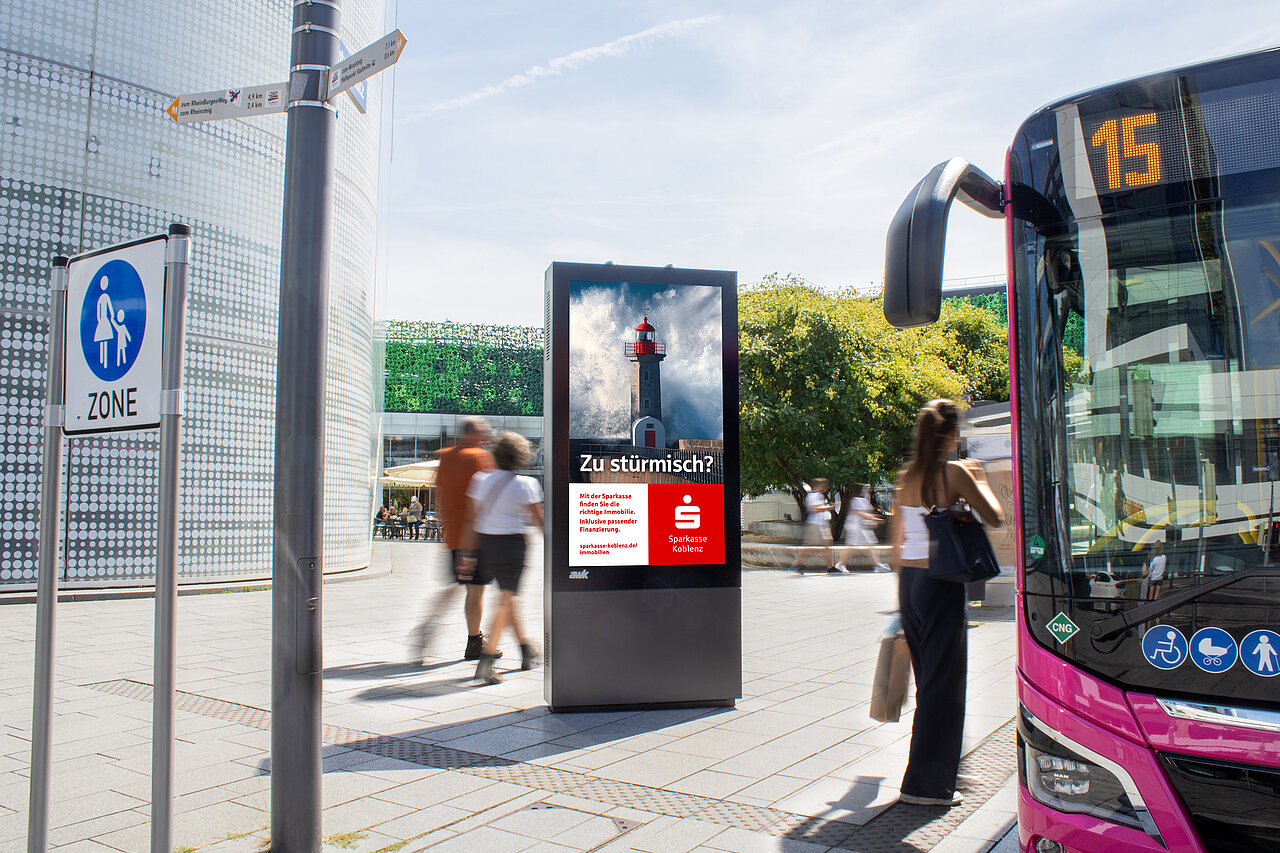 Digitale Vitrine mit Werbescreen an einer Bushaltestelle, vorbeigehenden Personen und einem haltenden Bus in einer belebten Stadtzone. Motiv von Sparkasse Koblenz und der Aufschrift: "Zu stürmisch?"