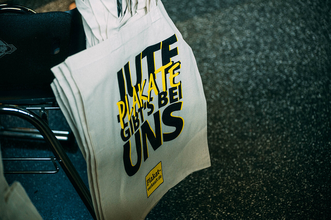 Jute-Beutel mit Aufdruck "Jute Plakate gibt's bei uns" und Markenlogo.