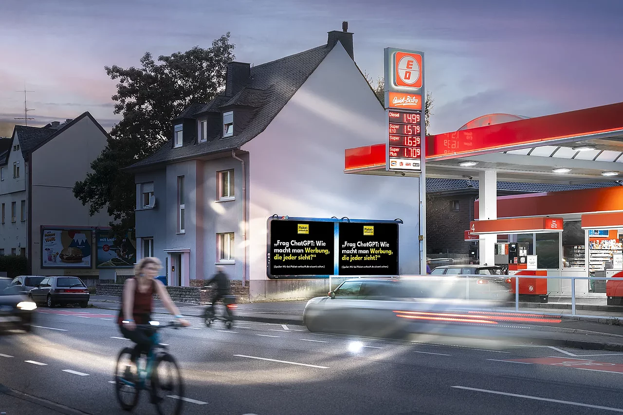 Großplakate mit der Aufschrift „Frag ChatGPT: Wie macht man Werbung die jeder sieht?" an einer Tankstelle mit vorbeifahrenden Autos und Radfahrern auf der Straße davor bei Sonnenuntergang.
