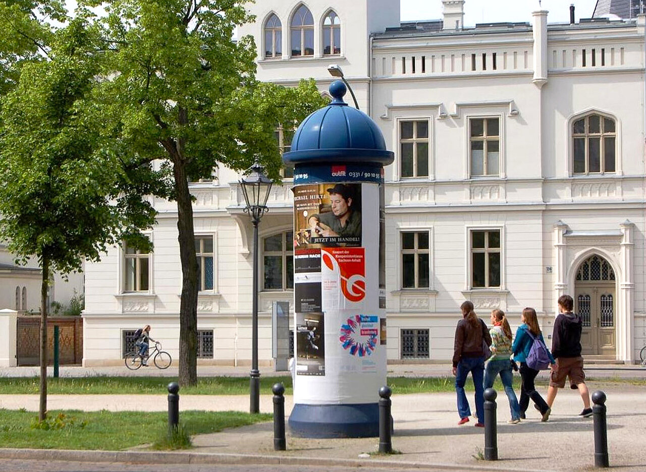 Werbesäule in Potsdam mit aktuellem Werbemotiv auf einem belebten Bürgersteig.