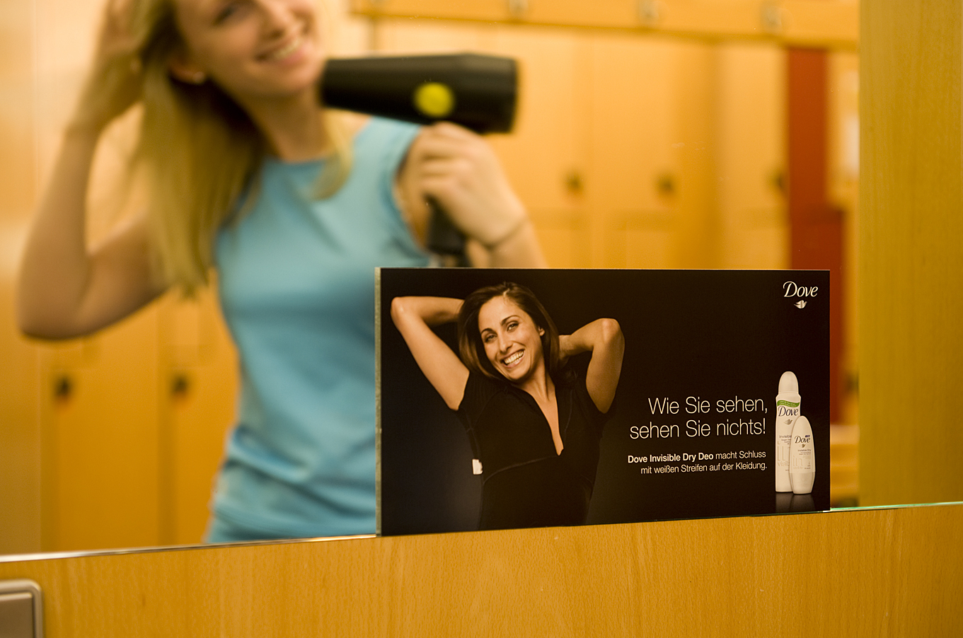 Werbeplakat von Dove, das auf einem Spiegel in einem Umkleidebereich angebracht ist, während im Hintergrund eine Frau ihre Haare föhnt – Beispiel für Spiegelwerbung in Sanitär- und Umkleideräumen.