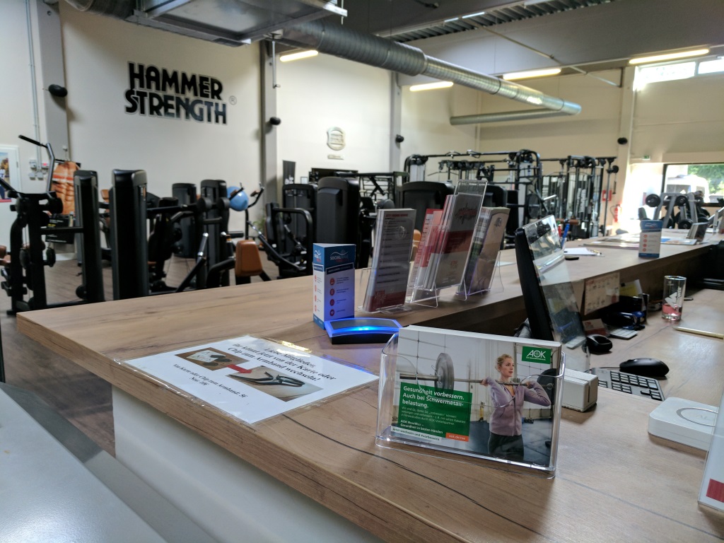 Thekenaufsteller mit Werbeflyern und Informationskarten auf einer Studiotheke in einem Fitnesscenter, im Hintergrund Trainingsgeräte und Hantelbereiche