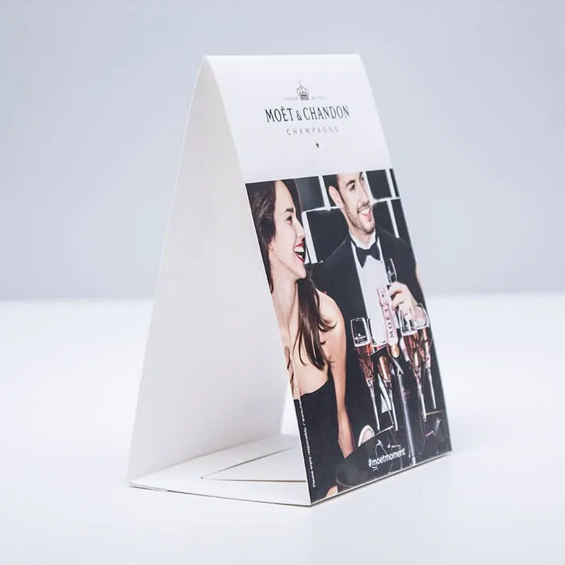 Eleganter Thekenaufsteller mit Moët & Chandon Werbung, aufgestellt auf einer weißen Fläche; zeigt ein festliches Motiv mit zwei feiernden Personen und Champagnergläsern.