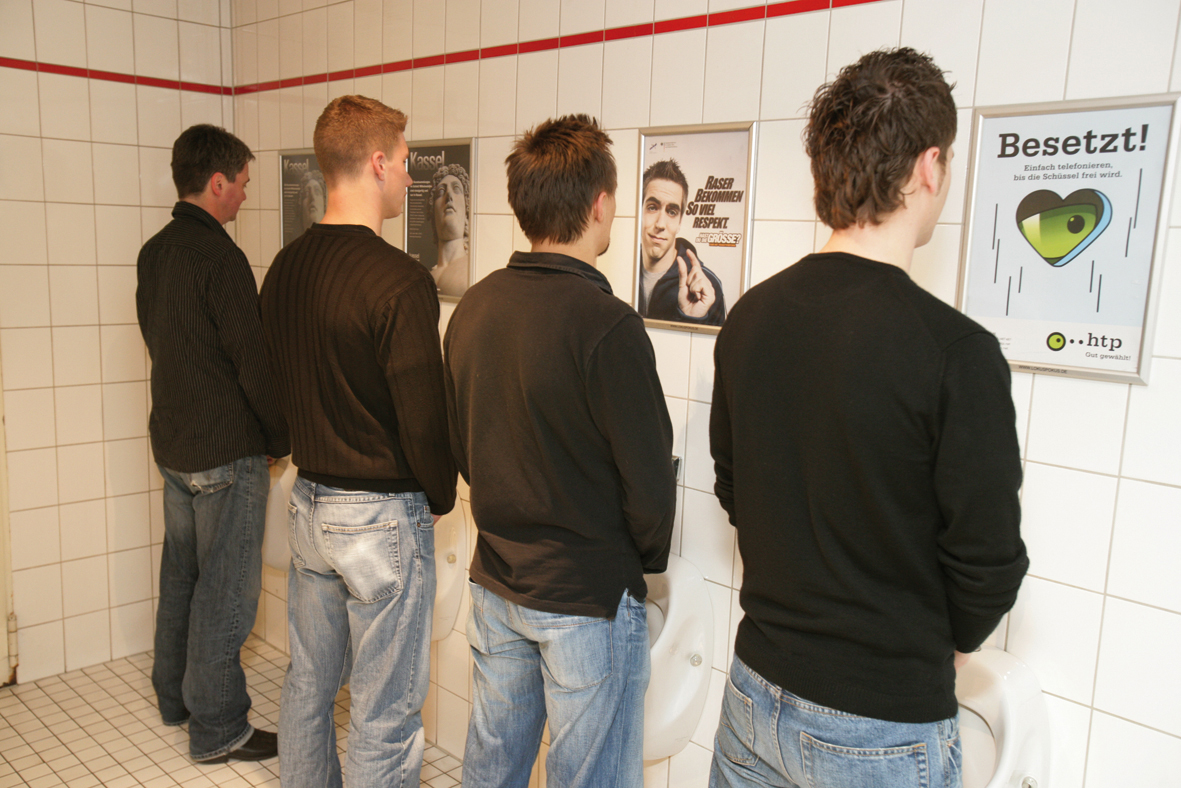Mehrere Personen stehen an Urinalen in einem Herrenwaschraum; direkt über den Urinalen hängen gut sichtbare Werbeplakate
